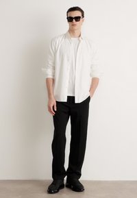 Vyras stovi prieš baltą sieną, vilkėdamas juodus saulės akinius, baltą marškinėlį su užtrauktuku virš baltos T-marškino, juodas kelnes ir juodus batus.