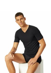 Camiseta negra de cuello en V en un tejido suave y elástico, combinada con pantalones cortos negros; modelo sentado en un pedestal blanco, fondo neutro.