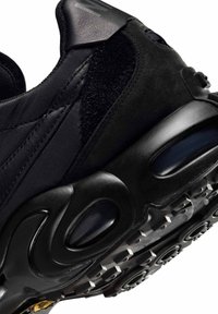 Primer plano de zapato deportivo negro que muestra malla texturizada, parte superior de ante y suela gruesa con patrón de tracción robusto y multidireccional.