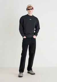 Svart sweatshirt med logotyp, svarta byxor och grå skor. Modellen bär solglasögon och en beige mössa. Enkel, avslappnad stil.