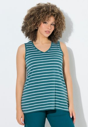 Woman with curly hair wearing a sleeveless teal and white striped top and teal pants, standing against a white background.