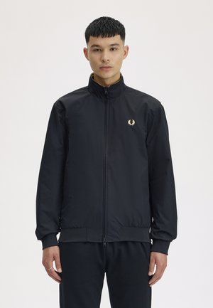 Ung mand iført en sort jakke med lynlås og et lille gyldent laurbærkranslogo på venstre bryst, stående mod en ensfarvet baggrund.