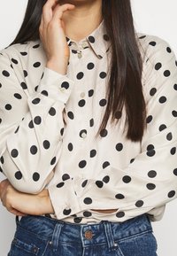 Camicia crema con bottoni e pois neri, dotata di colletto a punta e maniche lunghe. Leggero luccichio, con bottoni e polsini visibili.