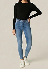 Personne portant un haut noir à manches longues, un jean skinny taille haute bleu clair et des baskets blanches, debout avec une main sur la hanche.
