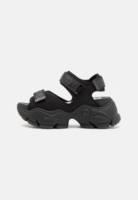 Sandales noires ouvertes à l'avant avec des semelles épaisses et texturées et des sangles ajustables. Fabriquées dans un matériau doux avec des accents perforés.