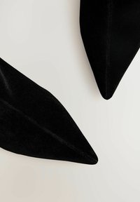 Czarne, aksamitne buty na szpilkach z ostrym czubkiem, charakteryzujące się gładką powierzchnią i gładką teksturą. Tło jest neutralne.