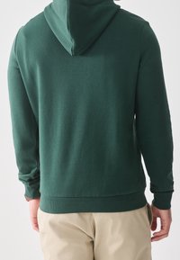 Uomo che indossa un felpa verde scuro e pantaloni beige, visto da dietro su uno sfondo chiaro e uniforme.