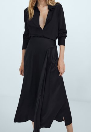 Femme portant un chemisier noir à manches longues avec un décolleté en V profond et une jupe midi noire avec un détail drapé et une fente latérale sur un fond uni.