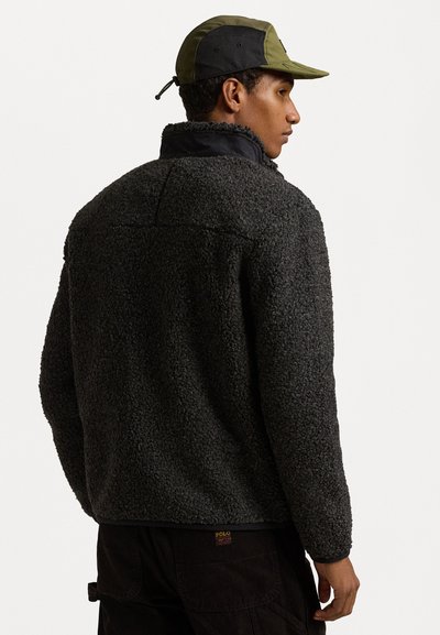 Fleece jakna v temno sivi barvi z teksturirano, puhasto zunanjostjo in visokim ovratnikom. Ujemajoča se dvobarvna kapa, ki prikazuje mrežno ploščo na zadnji strani.