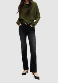 Olivgrüner Rollkragenpullover, lockere Passform; schwarze Jeans mit geradem Schnitt; schwarze, hochhackige Schuhe mit dekorativem Schnallendetail.
