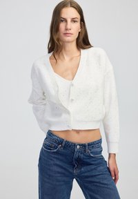 Kobieta w białym swetrze zapinanym na guziki z cekinami i pasującym topie, w zestawie z niebieskimi dżinsami o wysokim stanie, stojąca na jednolitym tle.