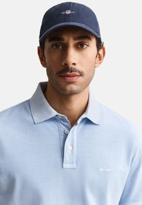 Mand med overskæg iført navyblå kasket og lyseblå poloshirt mod ensfarvet lyst baggrund, vendt fremad.