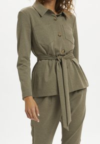 Olivgrünes, langärmliges Hemd mit Knopfleiste und Kragen, zwei Brusttaschen und einem Taillengürtel zum Hinzufügen einer Taille. Weicher, strukturierter Stoff.