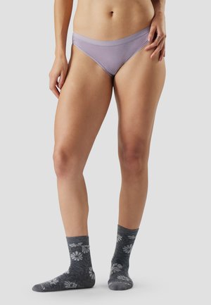 Vrouw met lichtpaarse onderkleding en grijze sokken met witte bloemenpatronen, staand tegen een effen lichte achtergrond.