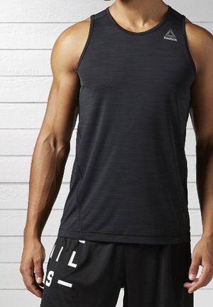 Lihaksikas henkilö, joka pukeutunut mustaan Reebok-merkkiseen hihattomaan urheilupaitaan ja mustiin shortseihin, joissa on valkoinen kirjainlogo, seisoo valkoista puuseinää vasten.