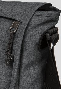 Šedá látková messenger taška s klopou, která má černý nastavitelný popruh a dekorativní detail s uzávěrem na přední straně.