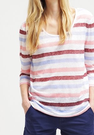 Kvinde iført en langærmet trøje med vandrette røde, pink og blå striber, hænderne i lommerne på marineblå bukser.