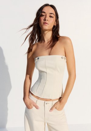 Jonge vrouw met lang donker haar draagt een strapless witte corsetachtige top en bijpassende hoge taillebroek, staand tegen een effen witte achtergrond.