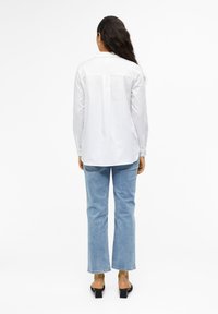 Camisa blanca de manga larga con un pliegue en la parte posterior, combinada con unos jeans acampanados de color azul claro. La modelo lleva unos zuecos de tacón negros. Textura de tela suave.