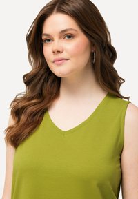 Top verde senza maniche con scollatura a V, realizzato in tessuto liscio. Presenta un design aderente e dettagli di cucitura minimi lungo il collo.
