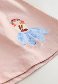 Pink cotton shirt featuring a printed design of a girl with red hair, blue flowers, and a blue tulle skirt. Close-up view of the hem.