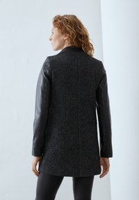 Cappotto nero a motivo houndstooth con maniche in pelle, caratterizzato da una vestibilità dritta e un colletto. La texture del tessuto appare spessa e calda.