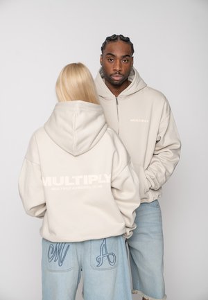 Two models wear light beige hoodies with "MULTIPLY" text; one faces forward, the other shows the back with brand logo, both in light denim bottoms.