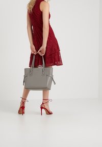 Femme dans une robe en dentelle rouge sans manches tenant un sac à main gris derrière son dos, portant des sandales à talons rouges avec des brides aux chevilles.