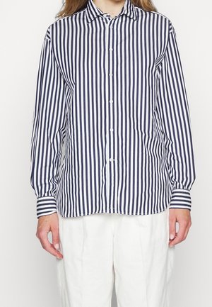 Personne portant une chemise à manches longues à rayures verticales noires et blanches, associée à un pantalon blanc, debout devant un fond uni.