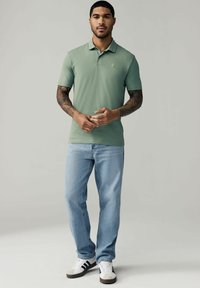 Polo de algodón verde con mangas cortas, combinado con jeans de color azul claro y zapatillas en blanco y negro. Presenta un pequeño logo bordado.