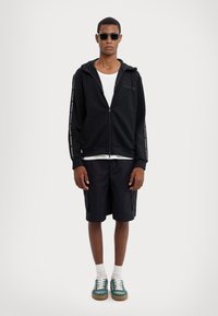 Sudadera negra con cremallera y logo, llevada sobre una camiseta blanca, combinada con pantalones cortos negros. Los complementos incluyen gafas de sol oscuras y zapatillas multicolores.