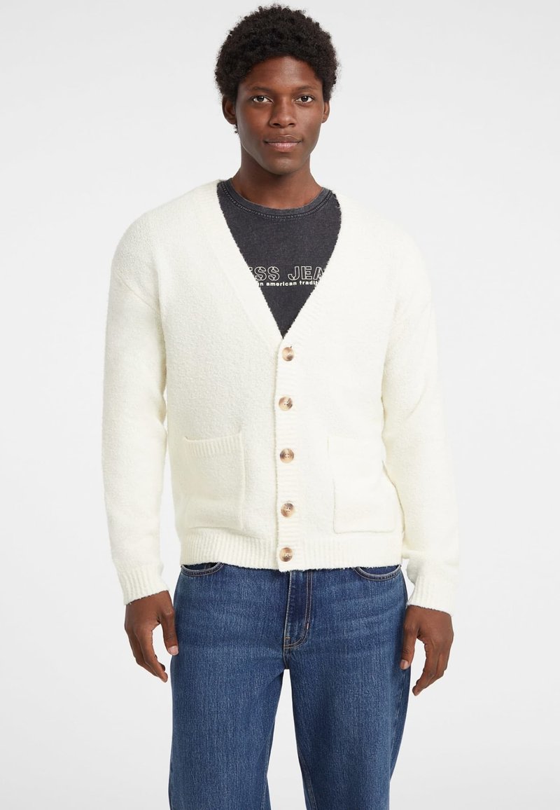 Witte, textuurgeweven cardigan met een diepe V-hals, houten knopen en twee zakken aan de voorkant, gedragen met denim jeans.