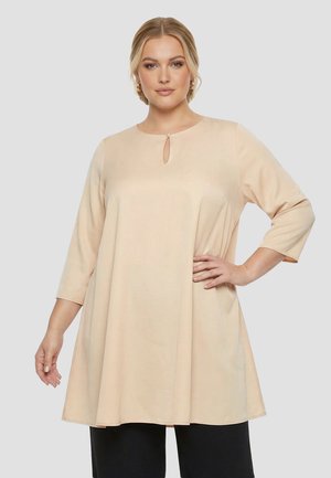 Kvinde iført en beige tunika med lange ærmer og en lille nøglehulning ved halsen, kombineret med sorte bukser, står med den ene hånd på hoften.