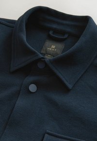 Col de chemise bleu marine foncé et boutons supérieurs avec une étiquette de marque noire visible indiquant "NEXT" taille medium.