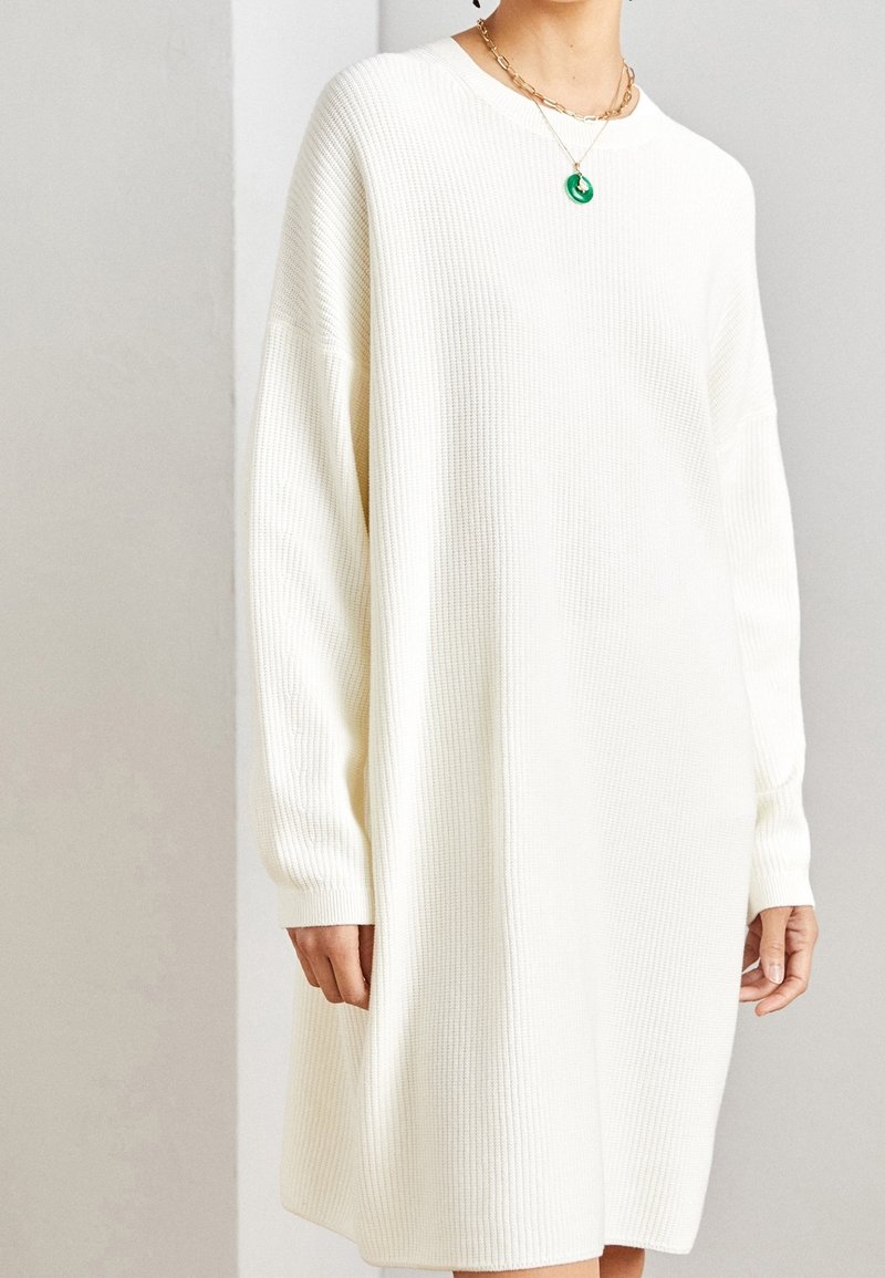 Robe à manches longues, oversize, en maille côtelée de couleur blanc cassé. Dotée d'un col rond, elle tombe au-dessus des genoux.
