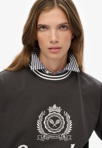 Dark gray sweatshirt featuring a white embroidered racquet club logo and a striped collared shirt underneath, with a ribbed neckline.