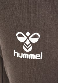 Brauner Stoff mit einem weißen Hummel-Logo in symmetrischem Design über dem Markennamen, was die Textur und den grafischen Kontrast betont.