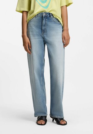 Person iført lyseblå vidde jeans, gul skjorte stoppet ind og sorte åbne tå hælede sandaler, står på hvid baggrund.