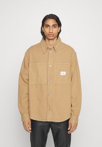 Camicia giacca oversize marrone in cotone con chiusura a bottone, due tasche sul petto e un'etichetta piccola sulla tasca sinistra.