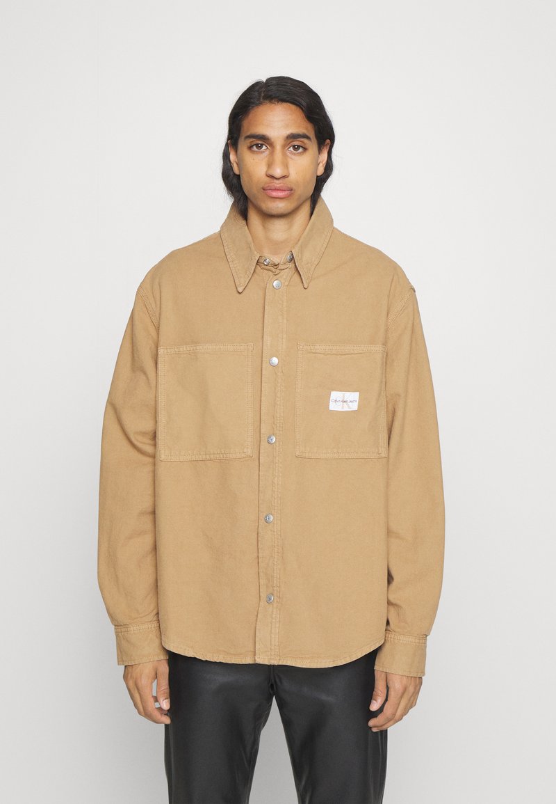 Camicia giacca oversize marrone in cotone con chiusura a bottone, due tasche sul petto e un'etichetta piccola sulla tasca sinistra.