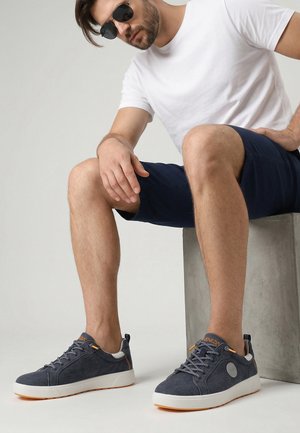 Man met een zonnebril, wit T-shirt, marineblauwe korte broek en grijze sneakers zittend op een grijze betonblok tegen een effen achtergrond.