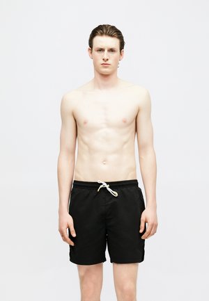 Jeune homme aux cheveux en arrière portant un short de bain noir avec un cordon blanc, debout devant un fond clair uni, les bras détendus.