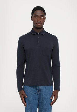 Mann iført en mørkeblå langermet polo med brystlomme og blå jeans, stående mot en ensfarget lys bakgrunn.