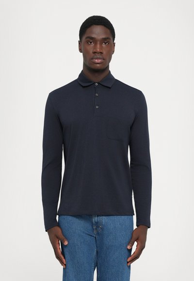 Homme portant un polo à manches longues bleu marine avec une poche poitrine et un jean bleu, debout contre un fond clair.