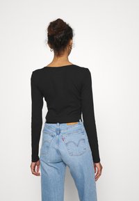 Nainen, jolla on nuttura ja kultaiset renkaat korvissa, pukeutuneena tiukkaan mustaan pitkähihaiseen paitaan ja korkeavyötäröisiin vaaleansinisiin farkkuihin, kääntää selkänsä.