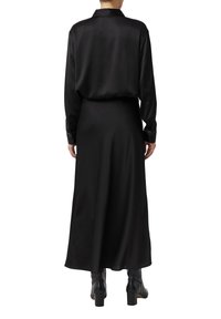 Osoba ubrana w długą czarną spódnicę, czarną bluzkę z długim rękawem i czarne botki na obcasie, stojąca plecami do widza.