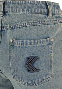 Los vaqueros de mezclilla azul claro presentan un bolsillo trasero con un logotipo oscuro bordado y remaches plateados. La tela tiene una textura clásica y un patrón de desgaste.