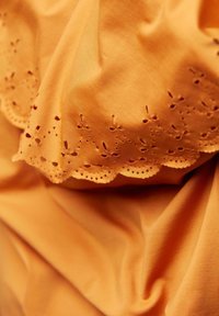 Baumwollstoff in lebhaftem Orange mit gewelltem Augenstickrand, der kleine Blumenmuster und dekorative Perforationen entlang des Randes aufweist.