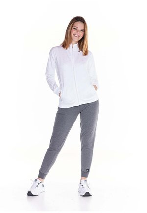 Une femme souriante porte un sweat à capuche blanc zippé avec des poches, un pantalon de jogging gris et des baskets blanches sur un fond blanc uni.