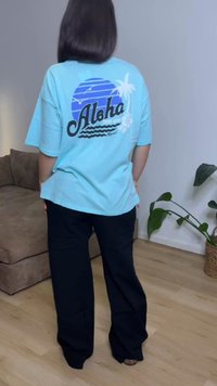 Άτομο που φοράει ανοιχτό μπλε φαρδιά μπλούζα με τη λέξη "Aloha" και τροπικό σχέδιο στην πλάτη, συνδυασμένη με μαύρο παντελόνι με φαρδιά πόδια σε εσωτερικό χώρο.